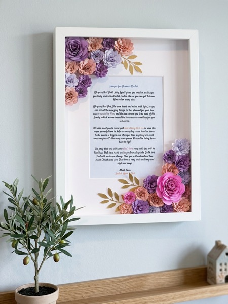 Tall white frame holding a written birthday prayer, with purple, peach and pink paper flowers in two corners.