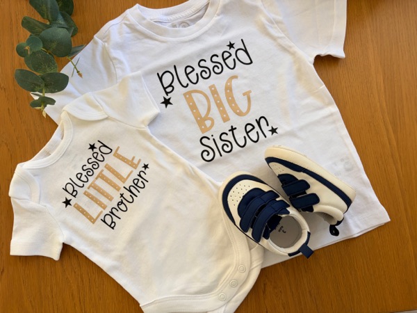 A white onesie lettered Blessed Little Brother beside a T-shirt lettered Blessed Big Sister, with baby shoes.